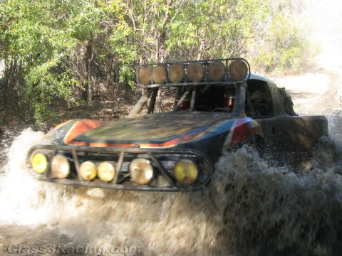 1990 Ford Bronco - off-road race truck topped with a roof light bar blasts through a watery section of dirt trail, kicking up brown spray.