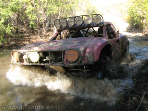 1990 Ford Bronco - faded, weathered off-road race truck numbered 16 with a roof light bar plows through a brushy river crossing in white spray.