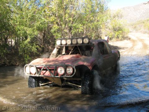 1990 Ford Bronco - battered red off-road race truck with a roof light bar and front light pod wades across a river crossing in a brushy canyon.