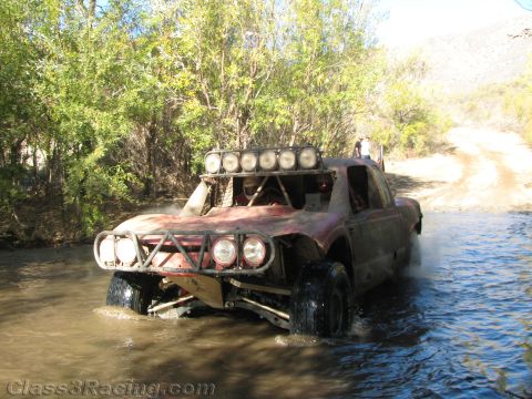 1990 Ford Bronco - weathered off-road race truck topped by a roof light bar fords a river crossing as spectators watch from the dirt trail behind.