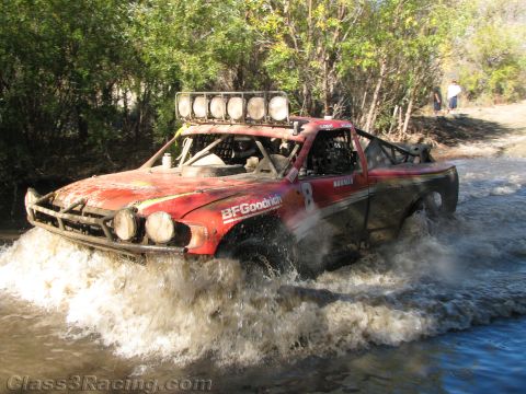 1990 Ford Bronco - red off-road race truck, number 8 with BFGoodrich livery and a roof light bar, charges through a river crossing in heavy spray.