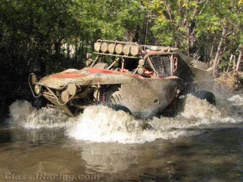 1990 Ford Bronco - heavily mud-caked open off-road race truck with a roof light bar plows through a deep river crossing, hurling water everywhere.