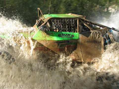 1990 Ford Bronco - green, mud-coated off-road race truck numbered 103 with Goodyear and King decals tears through a deep river crossing in a wall of spray.