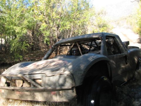 1990 Ford Bronco - heavily mud-coated off-road race buggy with a roll cage sits on a dirt trail surrounded by green brush.