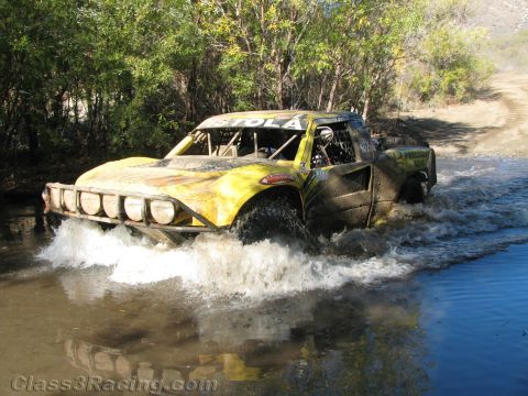 1990 Ford Bronco - yellow off-road race truck with light bar blasts through a shallow river crossing, throwing up a wide spray of water.
