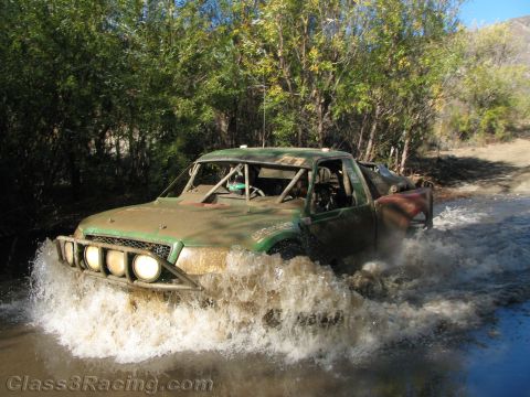 1990 Ford Bronco - green mud-splattered off-road race truck with front light pods churns through a stream on a wooded trail.