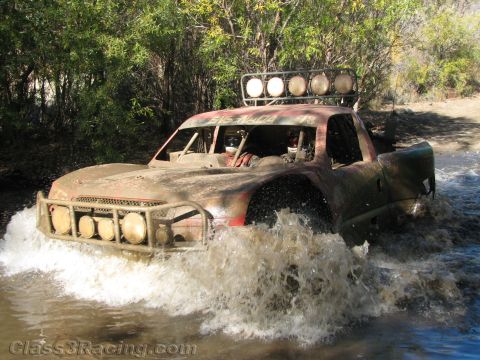 1990 Ford Bronco - muddy off-road race truck topped by a row of roof-mounted lights pushes through a shallow water crossing.