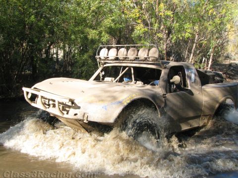 1990 Ford Bronco - tan, mud-encrusted off-road race truck with a roof light bar splashes through a creek beside green brush.