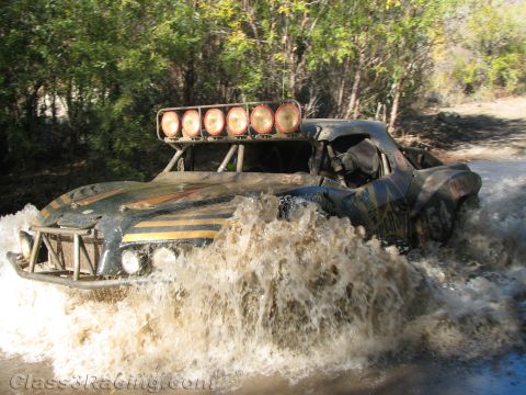 1990 Ford Bronco - dark off-road race buggy with six round roof-mounted lights blasts through a muddy water crossing.