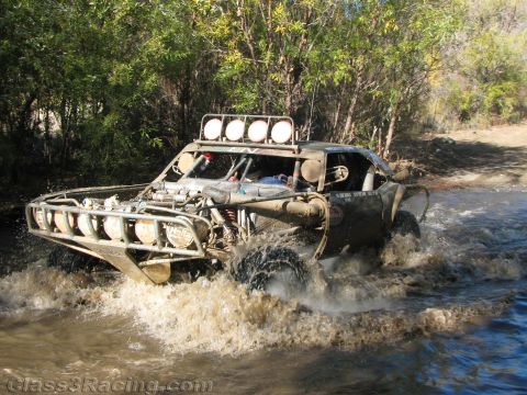1990 Ford Bronco - open off-road race buggy with exposed rear engine and front light pods crosses a muddy stream on a trail.