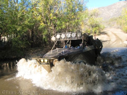 1990 Ford Bronco - off-road race vehicle with a five-light roof bar throws up a large brown splash crossing a river.