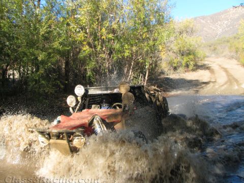 1990 Ford Bronco - off-road race buggy kicks up a huge spray of muddy water while crossing a stream on a wooded dirt trail.