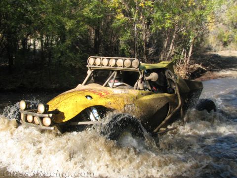1990 Ford Bronco - yellow off-road race buggy with a roof light bar and front pods crosses a river, spraying muddy water.