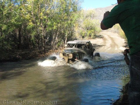 1990 Ford Bronco - small off-road buggy splashes across a shallow stream while a spectator in green stands at the right filming.