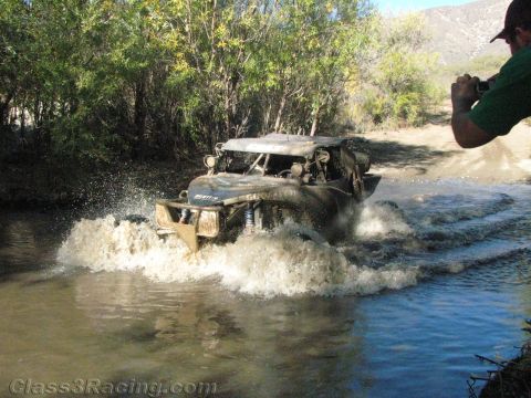 1990 Ford Bronco - off-road race buggy churns through a water crossing while a man in a cap photographs it from the bank.