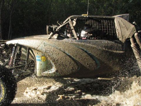 1990 Ford Bronco - close-up of a mud-spattered off-road race truck shows its netted cockpit and antenna as it runs through water.