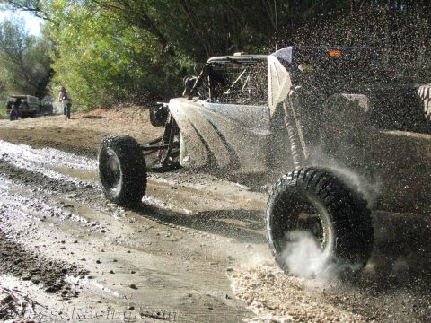 1990 Ford Bronco - open-wheel off-road race buggy throws up gravel and water spray while accelerating along a wet dirt road.