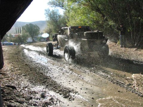1990 Ford Bronco - off-road buggy kicks up mud and spray racing down a wet rocky trail, with tents visible in the distance.