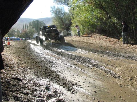 1990 Ford Bronco - off-road buggy churns along a muddy rutted trail while spectators and a photographer watch from the bank.
