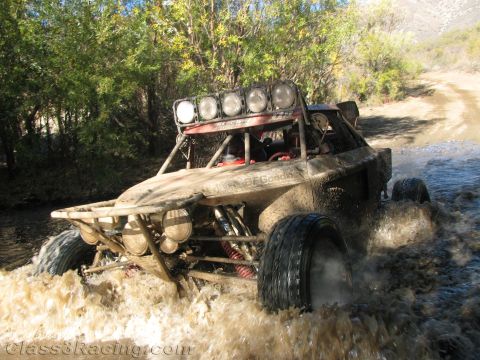 1990 Ford Bronco - off-road race buggy with four roof light pods splashes through a stream on a brush-lined dirt trail.