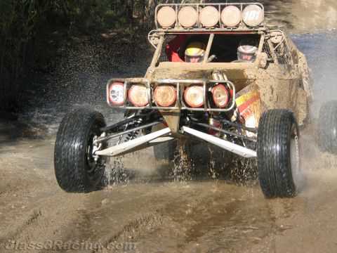 1990 Ford Bronco - off-road race buggy with a helmeted driver and round front lights launches through a shallow water crossing.