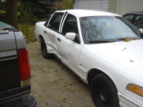 1997 Ford Crown Victoria - A white sedan shows extensive damage to its driver's side, with a dark pickup truck visible to its left.