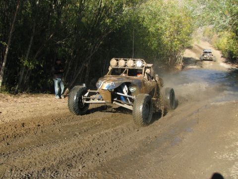 1990 Ford Bronco - off-road buggy with blue coilover shocks gets airborne on a muddy trail as a second buggy follows behind.