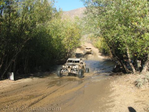 1990 Ford Bronco - Two off-road buggies make their way across a shallow stream along a green tree-lined dirt trail.