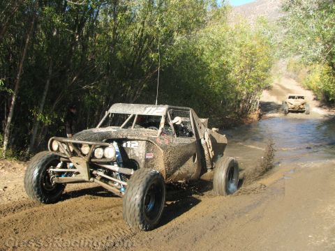 1990 Ford Bronco - mud-covered off-road buggy marked 112 runs along a wet dirt trail with a second buggy behind it.