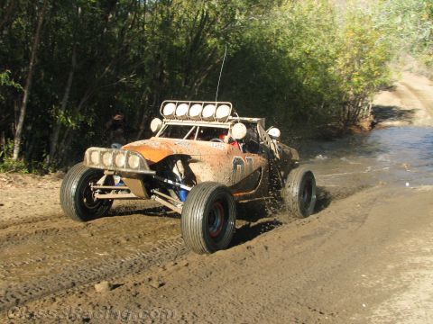 1990 Ford Bronco - orange single-seat off-road race buggy with a roof light bar churns through a muddy water crossing on a wooded dirt trail.