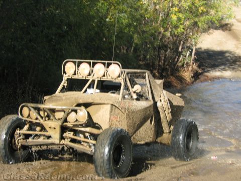 1990 Ford Bronco - heavily mud-covered off-road race buggy with a four-light roof bar and tall whip antenna powers through a shallow stream.