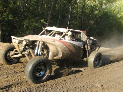 1990 Ford Bronco - long-travel off-road race buggy with exposed coilover shocks races along a dusty dirt course past brush.
