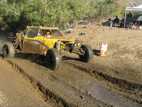 1990 Ford Bronco - yellow off-road race buggy crosses a deeply rutted muddy section near a pit area with tents and spectators in the background.