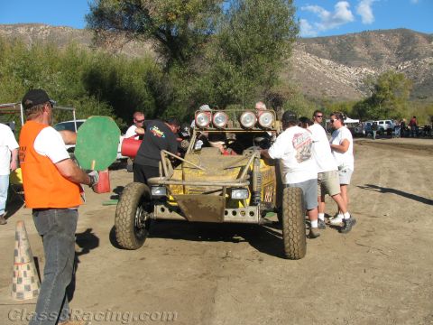 1990 Ford Bronco - yellow off-road race buggy sits front-on in a dirt pit as crew gather around and an official in an orange vest holds a sign.