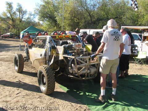 1990 Ford Bronco - numbered tube-frame off-road race buggy is serviced by crew at a dirt pit area laid with green mats under trees.