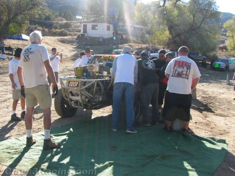 1990 Ford Bronco - group of crew members crowds around the back of a tube-frame off-road buggy on green pit mats below a hillside building.
