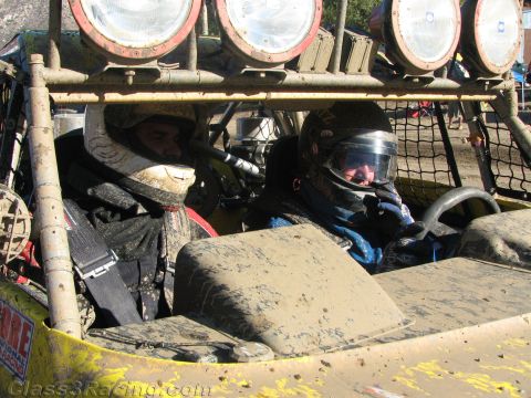 1990 Ford Bronco - Two helmeted occupants sit side by side in the muddy cockpit of an off-road race buggy beneath the roof light bar.