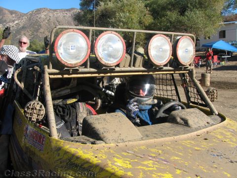 1990 Ford Bronco - rear of a worn yellow off-road buggy shows a four-light roof bar and a helmeted occupant in the cockpit at a pit area.