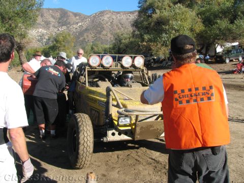 1990 Ford Bronco - Crew members push a yellow off-road buggy in a dirt pit while an official in an orange Checkers vest looks on, mountains behind.