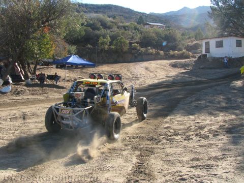 1990 Ford Bronco - yellow off-road race buggy kicks up dust on a dirt road below a hillside with a white camp building and tents.