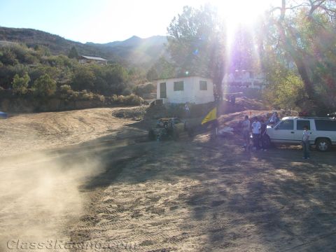 1990 Ford Bronco - off-road buggy raises dust in a sun-flared dirt clearing near a white building, with spectators and a pickup truck nearby.