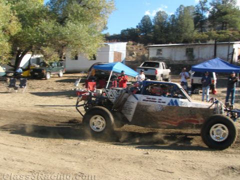 1990 Ford Bronco - silver and red tube-frame off-road race buggy accelerates across a dirt lot past pit canopies and parked pickups.