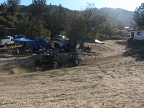 1990 Ford Bronco - off-road buggy drives across a sandy clearing toward camp tents and a hillside building under low afternoon sun.