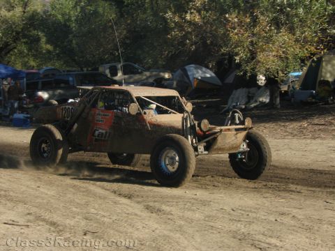 1990 Ford Bronco - tan tube-frame off-road race buggy speeds across a dirt area past camp tents and parked vehicles.