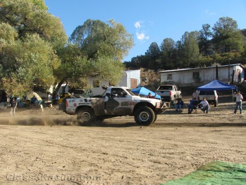 1990 Ford Bronco - white off-road race truck drives across a dirt lot, raising dust, below a hillside building and a blue pit canopy.