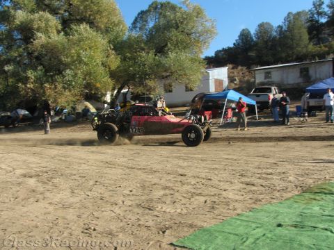 1990 Ford Bronco - pink and black off-road race buggy speeds across a dirt lot, throwing dust, near pit canopies and a parked truck.