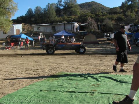 1990 Ford Bronco - orange and dark tube-frame off-road buggy crosses a dirt staging area as a man walks past, tents and spectators behind.