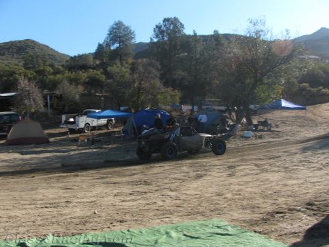 1990 Ford Bronco - off-road buggy drives along a sandy track past a campground of tents and canopies in a hilly desert valley.
