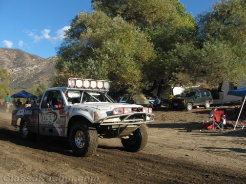 1990 Ford Bronco - white off-road race truck with a roof light bar drives along a dirt course past pit canopies and folding chairs.