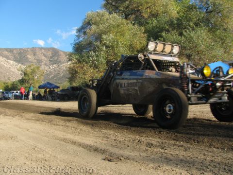 1990 Ford Bronco - dark off-road race buggy lettered 'VILLALOBOS' speeds along a dirt course beside a brushy hillside and pit area.